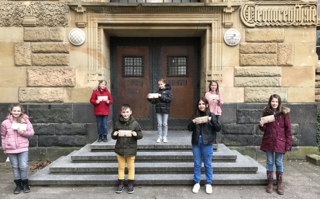 Ein Gruppenfoto der Schülerinnen und Schüler vor unserem Haupteingang mit einem Eulenpostumschlag in der Hand.