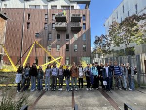 Ein Gruppenfoto der Schlüerinnen und Schüler in der Innenstadt vor einem rötlich-braunen Gebäude mit einem übergroßen gelben Maßband als Kunstinstallation im Hintergrund.
